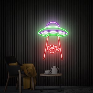 May include: A neon sign depicting a green and purple UFO with a red beam of light shining down on a cat. The sign is mounted on a dark, vertically striped wall. A black chair, a small table with a teapot and cup, and a yellow blanket are in the foreground.