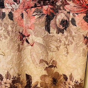 K&ouml;nnte beinhalten: Nahaufnahme eines floralen Stoffes mit strukturierter Oberfl&auml;che. Das Design zeigt gro&szlig;e, stilisierte Blumen in Rosa-, Rot- und Braunt&ouml;nen vor einem hellbeigen Hintergrund. Der Stoff hat einen subtilen Glanz, was auf eine formelle oder dekorative Verwendung hindeutet.