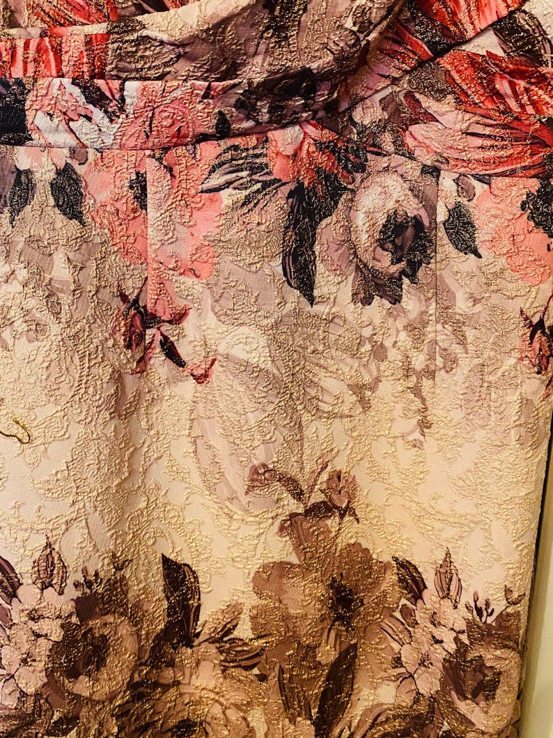 K&ouml;nnte beinhalten: Nahaufnahme eines floralen Stoffes mit strukturierter Oberfl&auml;che. Das Design zeigt gro&szlig;e, stilisierte Blumen in Rosa-, Rot- und Braunt&ouml;nen vor einem hellbeigen Hintergrund. Der Stoff hat einen subtilen Glanz, was auf eine formelle oder dekorative Verwendung hindeutet.