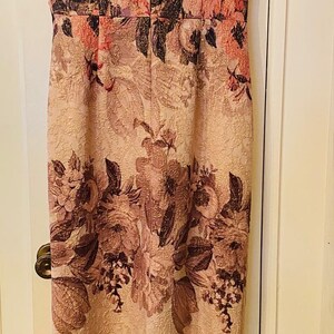 K&ouml;nnte beinhalten: &Auml;rmelloses, knielanges Kleid mit floralem Muster in Rosa-, Rot- und Braunt&ouml;nen. Das Kleid h&auml;ngt an einem wei&szlig;en Kleiderb&uuml;gel vor einer wei&szlig;en T&uuml;r. Das florale Design ist prominent.