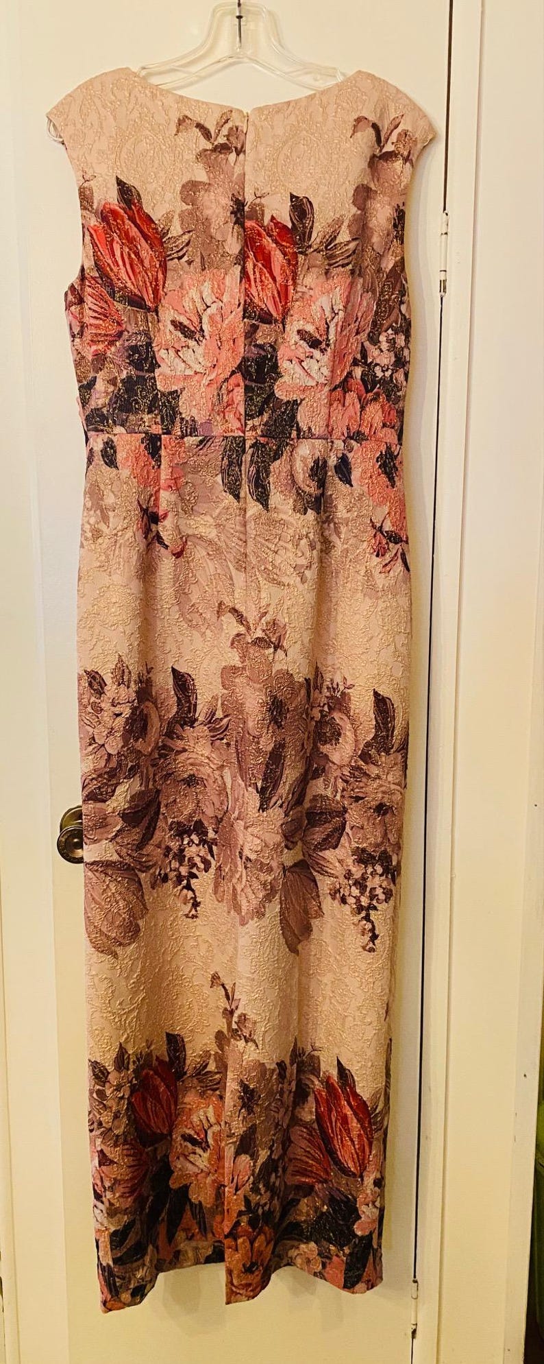 K&ouml;nnte beinhalten: &Auml;rmelloses, knielanges Kleid mit floralem Muster in Rosa-, Rot- und Braunt&ouml;nen. Das Kleid h&auml;ngt an einem wei&szlig;en Kleiderb&uuml;gel vor einer wei&szlig;en T&uuml;r. Das florale Design ist prominent.