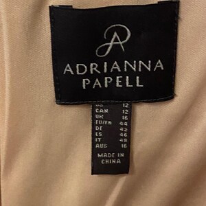 K&ouml;nnte beinhalten: Nahaufnahme eines schwarzen Etiketts mit dem Markennamen "ADRIANNA PAPELL" in wei&szlig;er Schrift. Das Etikett enth&auml;lt Gr&ouml;&szlig;enumrechnungen, einschlie&szlig;lich DE Gr&ouml;&szlig;e 42, und den Text "MADE IN CHINA". Der Hintergrund ist ein hellbeiger Stoff.