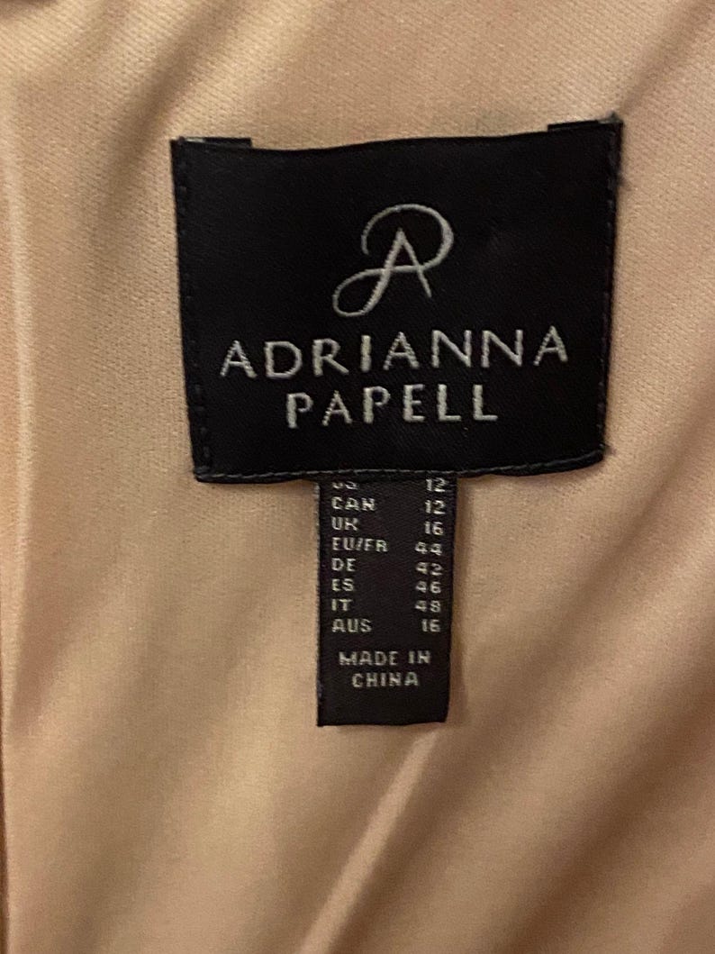K&ouml;nnte beinhalten: Nahaufnahme eines schwarzen Etiketts mit dem Markennamen "ADRIANNA PAPELL" in wei&szlig;er Schrift. Das Etikett enth&auml;lt Gr&ouml;&szlig;enumrechnungen, einschlie&szlig;lich DE Gr&ouml;&szlig;e 42, und den Text "MADE IN CHINA". Der Hintergrund ist ein hellbeiger Stoff.