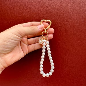 May include: A gold-colored heart-shaped keychain with a pearl beaded wristlet. The keychain has a lobster clasp and a small ring. The wristlet is made of white, round pearls. The background is a solid red color.