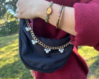 May include: A black crescent-shaped handbag with a gold chain and pearl accents. The bag is worn over the shoulder. A gold watch and a bracelet with colorful floral patterns are on the wrist.