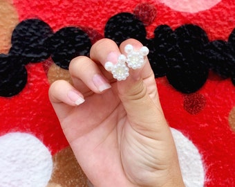 May include: A pair of white pearl earrings in the shape of a mouse. The earrings are held in a hand against a red background with black and white circular designs. The earrings have a central flower design.