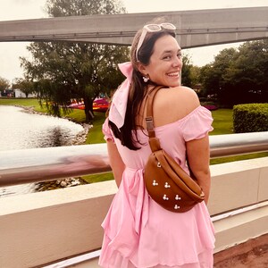 May include: A woman wearing a pink off-the-shoulder dress with a brown fanny pack with pearl accents. She is standing on a bridge with a body of water in the background.