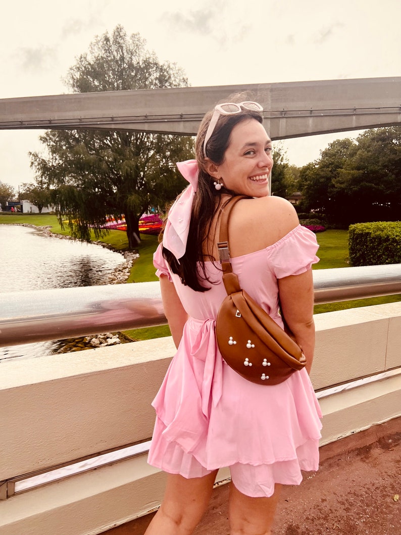 May include: A woman wearing a pink off-the-shoulder dress with a brown fanny pack with pearl accents. She is standing on a bridge with a body of water in the background.
