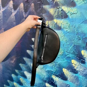 May include: A black leather fanny pack with a silver zipper and a silver buckle. The fanny pack is being held by a hand against a blue and yellow painted wall.