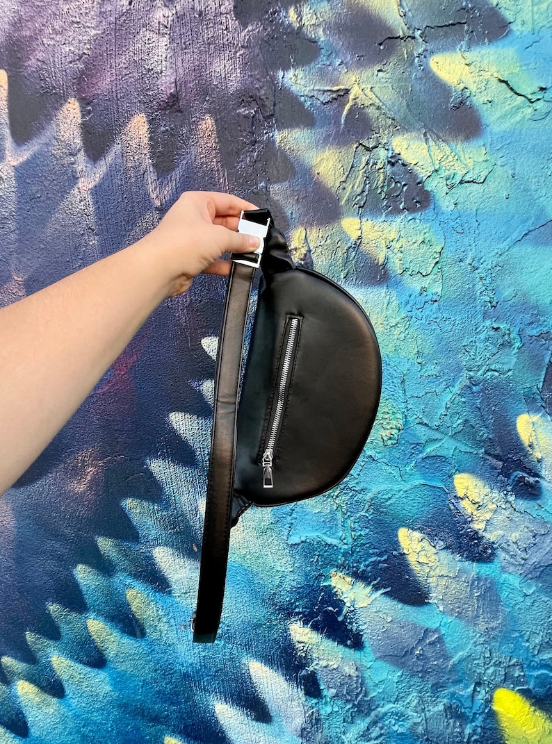 May include: A black leather fanny pack with a silver zipper and a silver buckle. The fanny pack is being held by a hand against a blue and yellow painted wall.