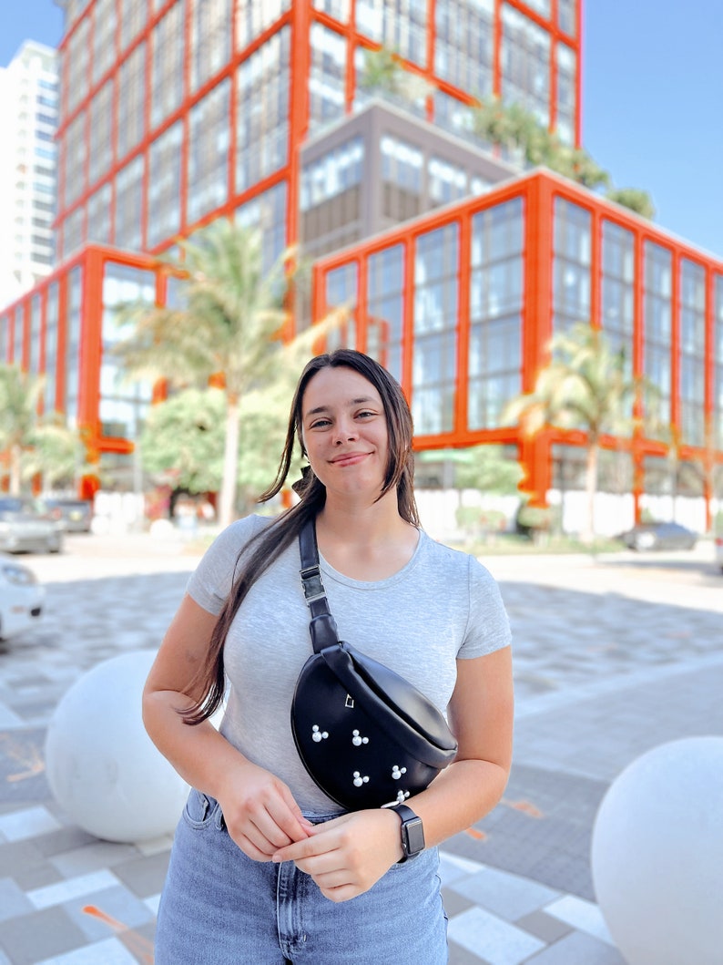 May include: A woman wearing a gray t-shirt and blue jeans is standing in front of a building. She is wearing a black fanny pack with pearl accents.