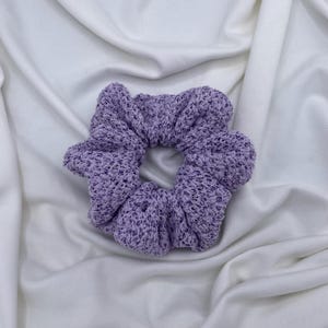 May include: A lavender scrunchie made of a textured knit fabric. The hair accessory is circular with a gathered design. The scrunchie is on a white fabric background.