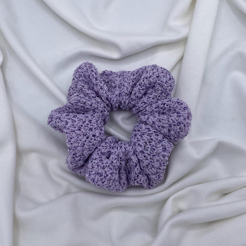May include: A lavender scrunchie made of a textured knit fabric. The hair accessory is circular with a gathered design. The scrunchie is on a white fabric background.