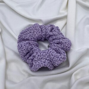 May include: A lavender scrunchie made of a knitted fabric. The hair accessory is circular and has a textured appearance. The scrunchie is set against a white, wrinkled fabric background.