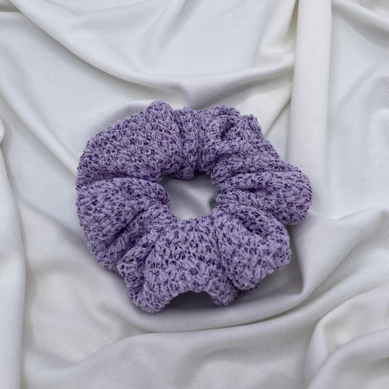 May include: A lavender scrunchie made of a knitted fabric. The hair accessory is circular and has a textured appearance. The scrunchie is set against a white, wrinkled fabric background.