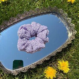 May include: A light purple scrunchie made of a crocheted material. The scrunchie is resting on a silver-colored, ornate, rectangular mirror. The mirror is surrounded by green grass and yellow flowers.