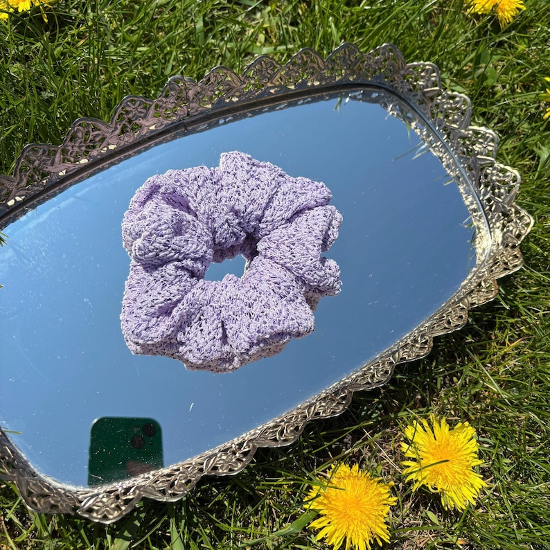 May include: A light purple scrunchie made of a crocheted material. The scrunchie is resting on a silver-colored, ornate, rectangular mirror. The mirror is surrounded by green grass and yellow flowers.