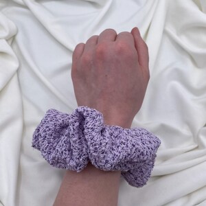May include: A light purple scrunchie made of a textured fabric, wrapped around a wrist. The scrunchie is a soft lavender color, with a slightly ruffled appearance. The background is a white, draped fabric.