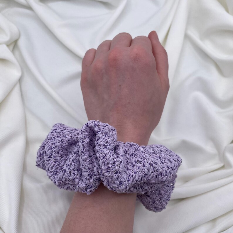 May include: A light purple scrunchie made of a textured fabric, wrapped around a wrist. The scrunchie is a soft lavender color, with a slightly ruffled appearance. The background is a white, draped fabric.