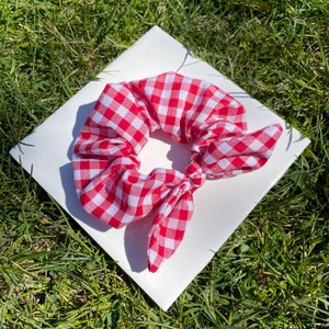 May include: Red and white gingham hair scrunchie with a bow. The scrunchie is on a white surface.