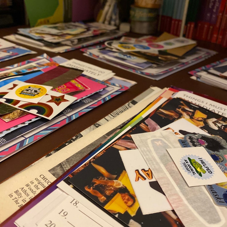 Op de afbeelding: Een close-up van een tafel bedekt met stapels kleurrijk papier, stickers en foto's. De tafel is bedekt met een bruine stof. De tekst "Things I must do immediately" is zichtbaar op een van de papieren.