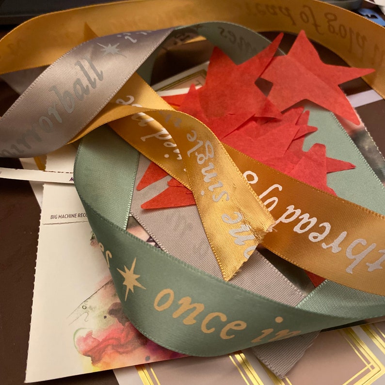 Op de afbeelding: Drie satijnen linten met gouden en zilveren letters. De linten zijn groen, goud en grijs. De tekst op de linten luidt "once in a blue moon", "the stars" en "the hall". Er zijn ook rode papieren sterren verspreid rond de linten.