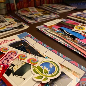 Op de afbeelding: Een close-up van een scrapbookpagina met een verscheidenheid aan kleurrijke stickers en foto's. De pagina is versierd met een blauwe en roze gepatenteerde rand. De stickers omvatten een tie-dye patroon, een hartontwerp en een wereldbol met groene bladeren.