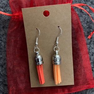 Peut inclure: Une paire de boucles d'oreilles en forme de pompon orange et rouge avec des crochets et des sommets argentés. Les boucles d'oreilles sont présentées sur une carte brune avec un trou en haut. La carte est dans un sac en maille rouge.