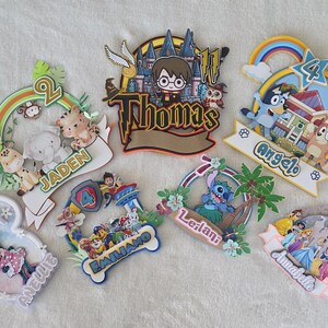 May include: A collection of seven multi-layered paper cake toppers featuring various cartoon characters, including Harry Potter with the name Thomas, Bluey with the name Angelo, Jaden, Emiliano, Leilani, Annabelle, and Minnie Mouse with the name Anellie.