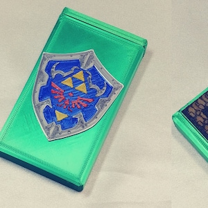 May include: Two 3D printed boxes. One box is green with a silver and blue shield with a red and yellow design. The other box is green with a black and brown patterned design. The text on the box reads "It's boring to go alone! Take these."