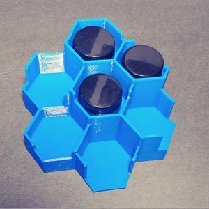May include: A blue hexagonal organizer with three compartments, each holding a black lidded jar. The organizer is made of plastic and has a honeycomb design.