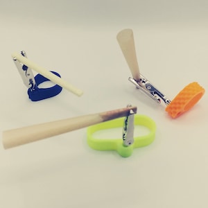May include: Three different colored plastic holders with metal clips for holding and securing items like incense sticks or rolled cigarettes. The holders are blue, green, and orange.