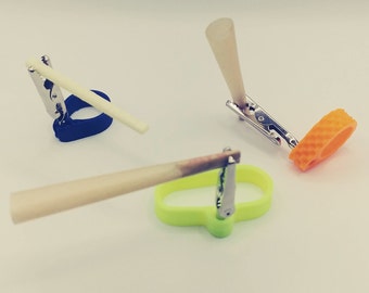 May include: Three different colored plastic holders with metal clips for holding and securing items like incense sticks or rolled cigarettes. The holders are blue, green, and orange.