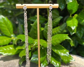 May include: A pair of silver dangle earrings with a single clear stone at the top of each earring. The earrings are hanging from a gold stand.