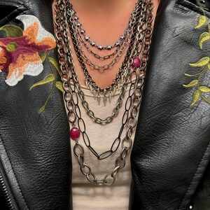 Può includere: Una collana a catena d'argento a più strati con più ciondoli, tra cui piccole punte e pietre rotonde rosa.