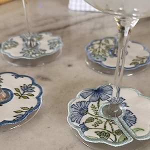 May include: Close-up of four floral embroidered drink coasters. Each coaster features a white fabric with blue and green floral embroidery, a light blue border, and a clear, round base. The coasters are designed to fit under wine glasses.