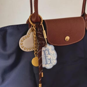 May include: A navy blue tote bag with brown leather handles and flap. A gold keyring holds a white heart charm with gold studs, a gold coin, a brown and white rope charm, and a blue floral tag with the letter "F".