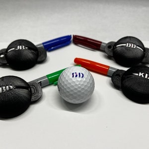 May include: A selection of black 3D-printed golf ball markers, each with initials, surrounding a golf ball. The markers are displayed with coloured Sharpie pens. The golf ball also has initials printed on it.