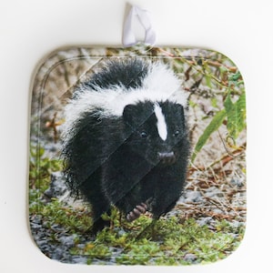 May include: A black and white skunk pot holder with a quilted design. The skunk is walking through a green grassy area.