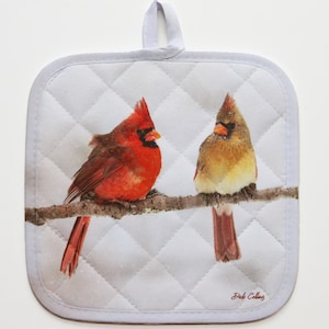 May include: White quilted pot holder with a red cardinal and a brown cardinal perched on a branch. The pot holder has a loop for hanging. The text "Dick Collins" is printed in the bottom right corner.