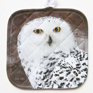 May include: A brown quilted hot pad with a snowy owl image. The owl has yellow eyes and white and black feathers.