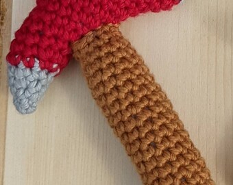 Pode incluir: Um machado de brinquedo de crochê feito à mão. A cabeça do machado é vermelha com uma borda cinza e o cabo é marrom. O brinquedo é feito de fio macio e é adequado para crianças. O brinquedo é colocado em uma superfície de madeira clara.
