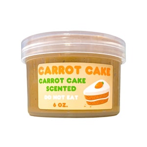 Könnte beinhalten: Ein durchsichtiges Plastikglas gefüllt mit einer braunen, cremigen Substanz. Das Glas hat ein Etikett mit der Aufschrift "Carrot Cake Carrot Cake Scented Do Not Eat 6 oz.". Das Etikett zeigt auch ein Cartoon-Bild eines Stückes Karottenkuchen mit einer Karotte oben drauf.