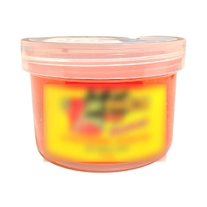 Könnte beinhalten: Ein kleines, klares Plastikglas gefüllt mit einer leuchtend orangen Substanz. Das Glas hat ein gelbes Etikett mit schwarzem Text, der verschwommen ist.