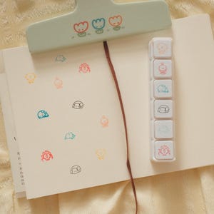 May include: A white notebook with a light green clip featuring three small tulip illustrations. The notebook contains various small, colorful animal stamps and includes a vertical white stamp set with six distinct icons and a thin brown bookmark ribbon.