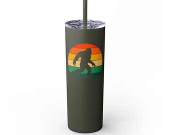 May include: A green stainless steel tumbler with a retro-style graphic of a Bigfoot walking in front of a sunset. The tumbler has a lid and a straw.