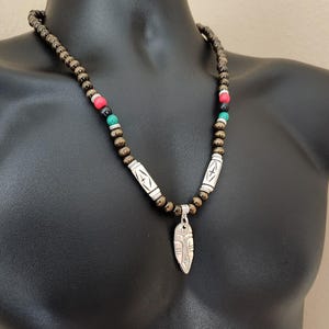 May include: A beaded necklace with a silver pendant. The necklace features dark brown beads, interspersed with red, green, and white beads. The pendant is a silver, shield-shaped design. The necklace is displayed on a black mannequin.