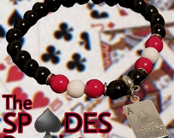 May include: Black beaded bracelet with red and white beads and a silver charm of the Ace of Spades card. The bracelet is on a background of playing cards. The text "The SPADES Bracelet exclusively @sankofa.wear" is at the bottom of the image.