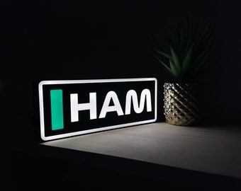 May include: A black and white sign with the word "HAM" in white letters and a green vertical bar to the left of the word. The sign is illuminated and sitting on a light wood surface.