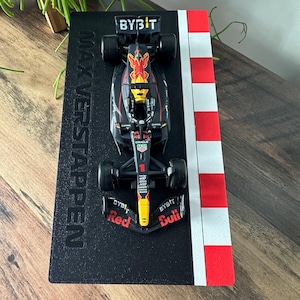 May include: A black and yellow Formula 1 race car model with the words "BYBIT" and "Red Bull" on the side. The car is on a black display stand with the words "MAX VERSTAPPEN" printed on it. The stand is next to a red and white chequered race track.